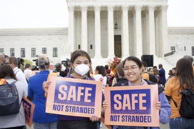 Image of two people at a rally in front of the Supreme Court holding signs that say "Safe Not Stranded"